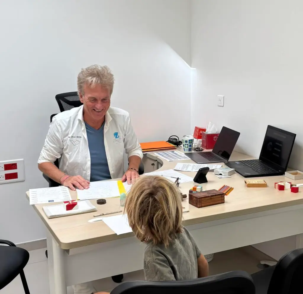 Consultation avec un enfant vu de dos, autour d'un bureau couvert de tests, de jeux et d'ordinateurs