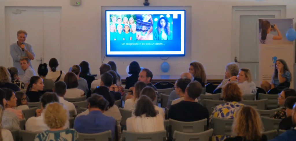Conférence devant une salle pleine : l'intervenant debout au micro à gauche de l'écran